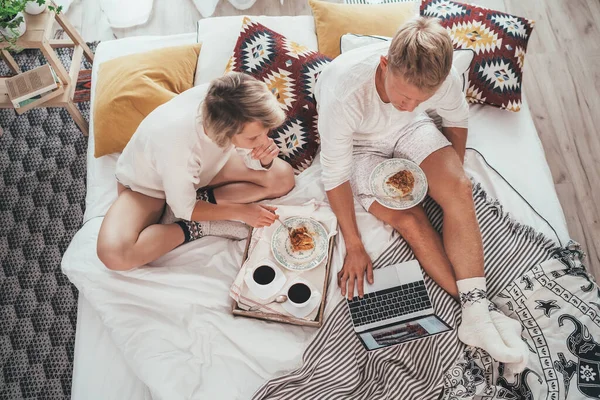 居心地の良いベッドでパジャマでカップル一緒にラップトップを使用してインターネットを閲覧し アップルパイデザートと朝のコーヒーを持っています — ストック写真