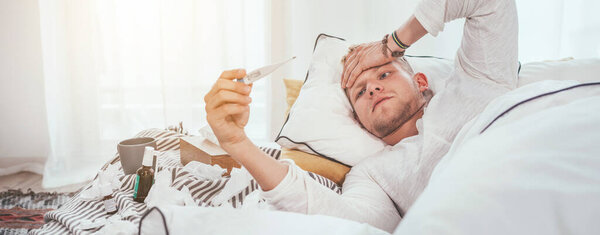 Unhealthy and tired looking at a medical thermometer as lying in a cozy home bed beside a lot of used paper tissues and medicines on the blanket. Season virus flu and a home quarantine concept image.