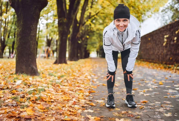 若い運動に適した笑顔の女性は 秋の都市公園でジョギングした後 足の途中で膝の上に立って 最新のランニング服を着ていました 実行中の人のコンセプトイメージ — ストック写真
