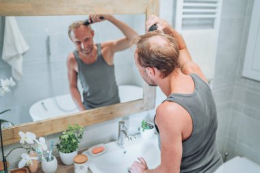 Vücut ve cilt bakımı. Gülümseyen adam banyo aynasında kendine yeni bir saç kesimi yapıyor. Banyodaki aynaya bakıyor. Kişisel bakım, saç stili, kendine özen, erkek güzelliği.