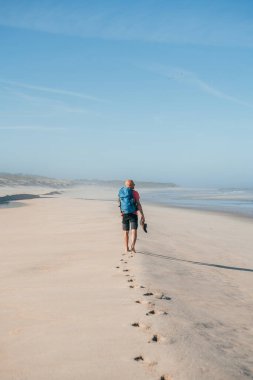 Yalnız, çıplak ayaklı, sırt çantalı bir adam sabahın köründe Atlantik Okyanusu kıyısında kumlu Portekiz sahilinde yürüyor. Camino Portekiz 'de çekilen popüler hac güzergahı. Aktif insanlar, seyahat konsepti.