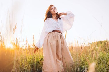 Beyaz bluz ve bej etekli genç bir kadın altın çayır boyunca özgürce yürüyor günbatımının keyfini çıkarıyor sıcak güneş ışığının barışçıl doğanın atmosferinin tadını çıkarıyor özgürlüğün farkındalığını ve doğayla uyum içinde olduğunu ifade ediyor.