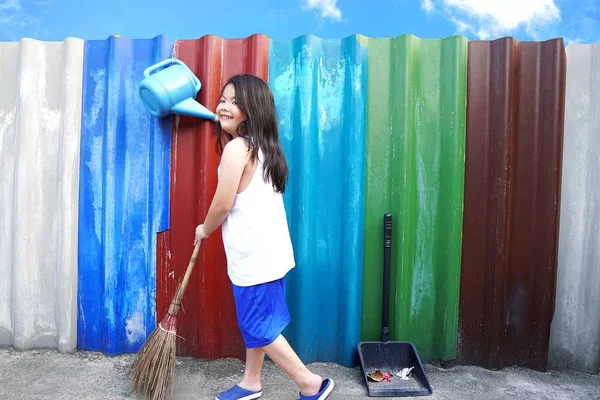 Asian little cute girl sweeping the floor with broom at outdoor on colourful wall and blue sky background.Inspired kid doing household chore.People,Housework, cleaning and housekeeping concept.