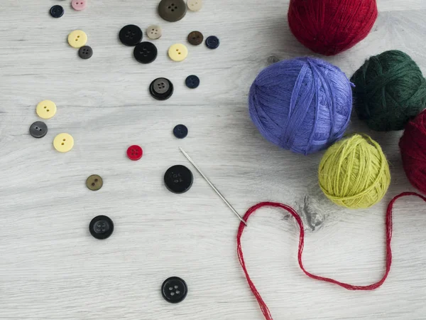 All for needlework: Buttons, a needle with a red thread, tangles of colored threads on a wooden gray table. Top view — Stock Photo, Image