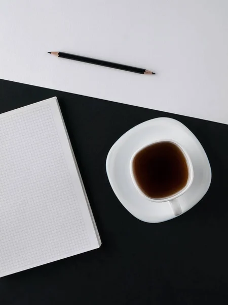 Office concept. Black pencil, blank notebook and white cup with coffee on black and white paper background. Top view. Copy space