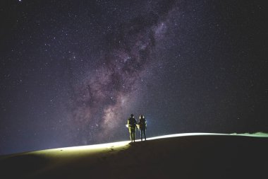 Samanyolu, yatay. Gece gökyüzünde yıldızlar ve kum tepe üzerinde duran birkaç silueti ile.