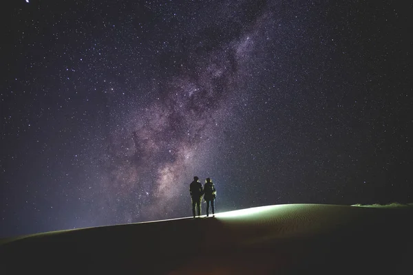 Samanyolu, yatay. Gece gökyüzünde yıldızlar ve kum tepe üzerinde duran birkaç silueti ile.