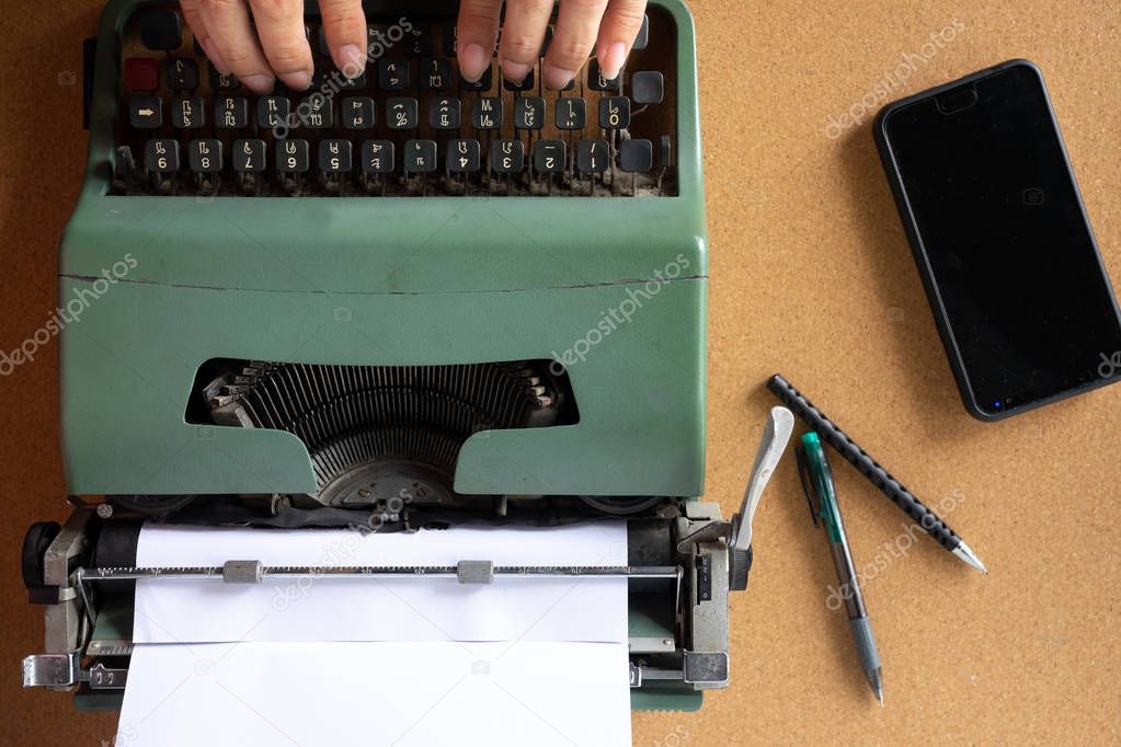 Two man hand typing on Vintage old mint green Thai typewriter with plain white paper on cork wooden board with pens and mobile phone on side.