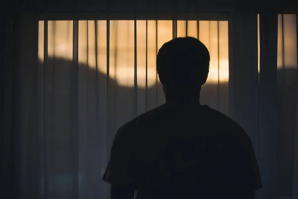 Back view of sad man standing in front of curtain and looking out window