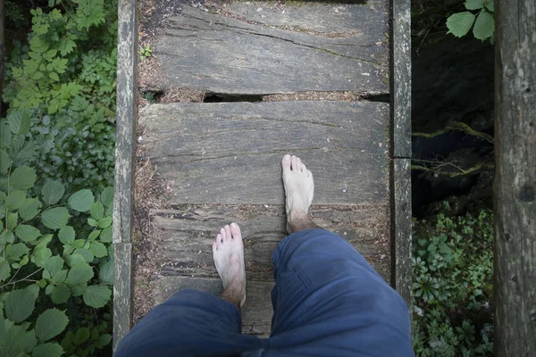 Barefoot man crossing old damaged wooden bridge in forest