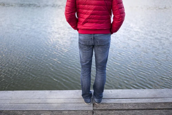 Thoughtful young man in jeans and red jacket stands on the edge of a wooden pier and observing blurry lake. Selective focus used.