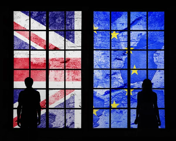 Silhouette of a couple standing and looking through big window at cracked and damaged United Kingdom and European union flags.
