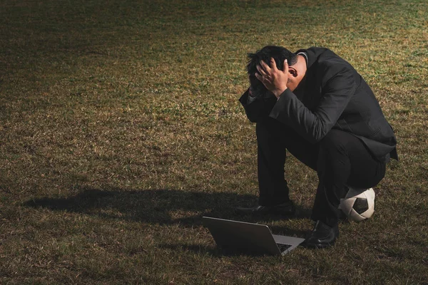 Stressed businessman with head in hands at soccer field, Stressed young businessman sitting outside corporate office working on laptop computer holding head, Business failure concept