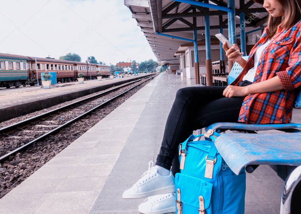 Young woman traveler and backpack waiting railway at train station, Young woman sitting with using smartphone at the train station, Summer holiday and travel concept, Sun light flare