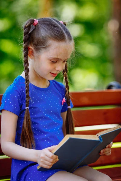 Portret van schoolmeisje op een bankje lezen boek. Achtergrond-stadspark. — Stockfoto