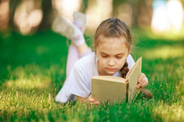 Küçük kız yeşil çimlerde yatar ve Yeşil Park 'ta kitap okur..