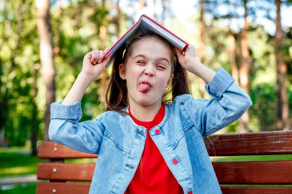 ベンチの上の公園の女の子は頭に本を持って顔を作る — ストック写真