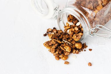 Bir kavanoz ev yapımı granola beyaz bir masanın üstüne döküldü. Bu granola lezzetli bir kahvaltı için çikolatalı, tarçınlı ve bademli kızarmış yulaftan yapılır. Boşluğu kopyala.