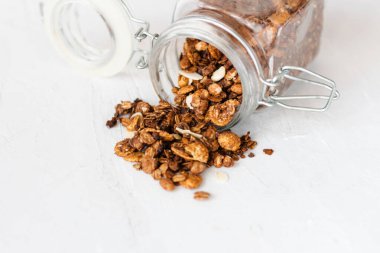 Bir kavanoz ev yapımı granola beyaz bir masanın üstüne döküldü. Bu granola lezzetli bir kahvaltı için çikolatalı, tarçınlı ve bademli kızarmış yulaftan yapılır. Boşluğu kopyala.