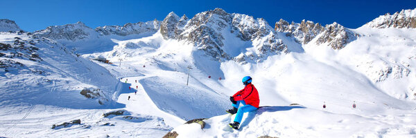 Молодой счастливый лыжник, сидящий на вершине гор и наслаждающийся видом на Ретийские Альпы, перевал Тонале, Италия, Европа
.  