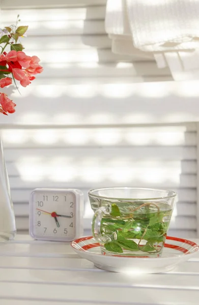 Bir saat, çiçek ve güneş ışınları ile beyaz bir arka plan üzerinde bir cam fincan nane çayı.