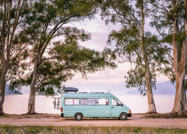 Camper van Park ve sakin ve huzurlu deniz içinde belgili tanımlık geçmiş ile akşam ışık, güzel uzun boylu ağaçlar arasında çerçeveli.