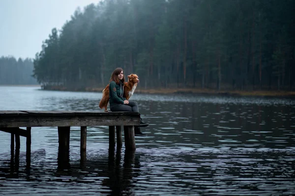 Bir köpek ile kız gölde bir iskelede oturup. Nova Scotia Duck Tolling Retriever doğa üzerinde bir adam ile — Stok fotoğraf