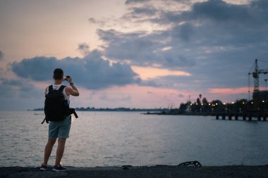 Bir adam gün batımını ve denizi fotoğraflıyor. Kumsalda sırt çantalı bir adam gün batımında akıllı bir telefonla denizin fotoğraflarını çekiyor.