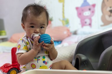 Bebek kız yumuşak oyuncaklarla çevrili bir yatakta oturuyor, elinde bir oyuncakla oynuyor.