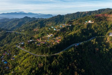 Güneşin doğuşuyla yemyeşil bir araziye uzanan dolambaçlı bir dağ yolunun hava manzarası.