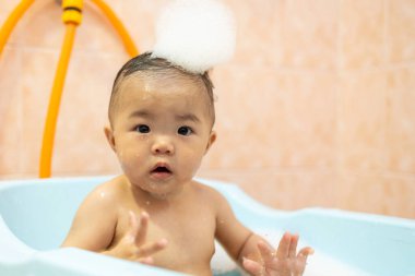Güzel Asyalı bebek, küvette oturmuş sabun köpüğüyle banyo yapıyor.