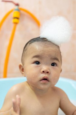 Güzel Asyalı bebek, küvette oturmuş sabun köpüğüyle banyo yapıyor.