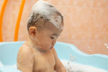 Güzel Asyalı bebek, küvette oturmuş sabun köpüğüyle banyo yapıyor.