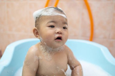 Güzel Asyalı bebek, küvette oturmuş sabun köpüğüyle banyo yapıyor.