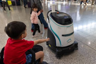 Hong Kong, Çin - 21 Mart 2024 - Bir kız ve babası arka planda yürürken genç bir çocuk bir robot temizleyiciye dokunuyor