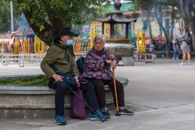 Hong Kong, Çin - 23 Mart 2024: Koruyucu yüz maskesi takan yaşlı çift, bir bankta oturuyor ve dini bir tapınağı ziyaret ettikten sonra dinleniyor