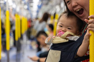 Anne, gülümseyen bebeğini bir taşıyıcıda tutarken trene biniyor.