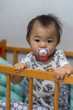 Sevimli Asyalı bebek beşikte dururken renkli bir emzik çiğniyor.