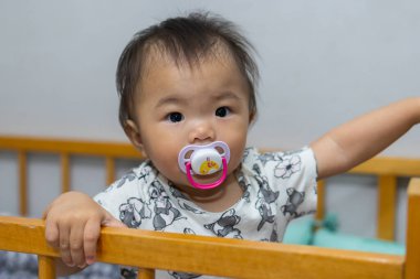 Sevimli Asyalı bebek beşikte dururken renkli bir emzik çiğniyor.