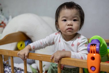 Sevimli bir kız bebek beşikte duruyor, korkuluklara tutunuyor, ileriye bakıyor.