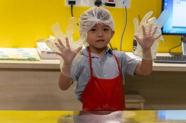 Asyalı çocuk saç filesi ve kırmızı önlük takıyor. Mutfakta tek kullanımlık eldiven gösteriyor.