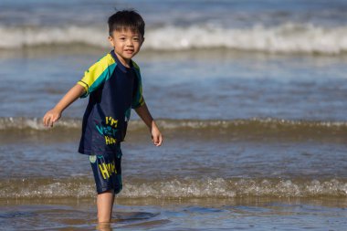 Plaj günü eğlencesi: Renkli bir dinozor temalı bebek tulumu giymiş bir çocuk kıyıda yürüyor, kumlara ve küçük bir küreğe odaklanıyor.