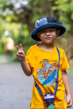 Şapkalı ve su şişeli Asyalı çocuk tropikal bir parkta yürürken zafer jesti yapıyor.