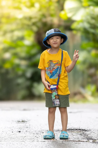 Şapkalı ve su şişeli Asyalı çocuk tropikal bir parkta yürürken zafer jesti yapıyor.