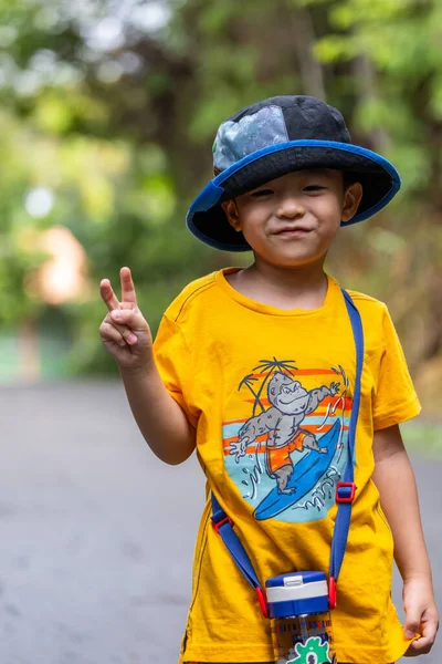 Şapkalı ve su şişeli Asyalı çocuk tropikal bir parkta yürürken zafer jesti yapıyor.