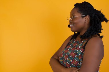 Sarı arka planda gözlüklü, neşeli, güzel, Afro-Amerikan bir kadının portresi. Kollarını kavuşturup güzel bir sohbet yapıyor.