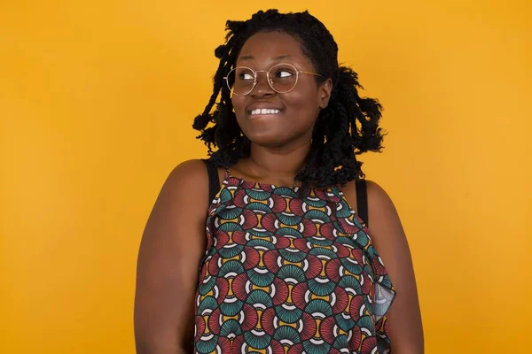 Photo of amazed  Young beautiful afro american woman wearing glasses over yellow background  bitting lip and looking up to empty space,