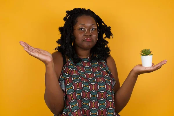 Hesitant Young beautiful afro american woman wearing glasses over yellow background  shrugs shoulders, looks uncertain and confused. Have no answer