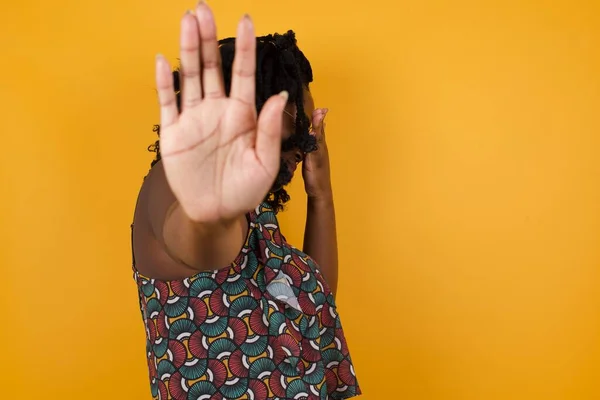 People, body language. Young beautiful afro american woman wearing glasses over yellow background  covers eyes with palm and doing stop gesture, tries to hide from everybody.