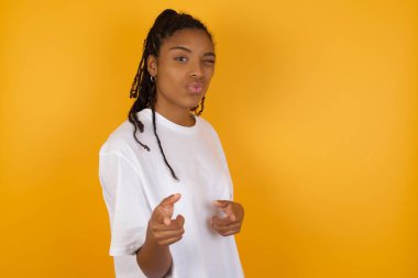 Afro-Amerikan kadın göz kırpıyor ve sarı arka planda kameraya işaret ediyor. 